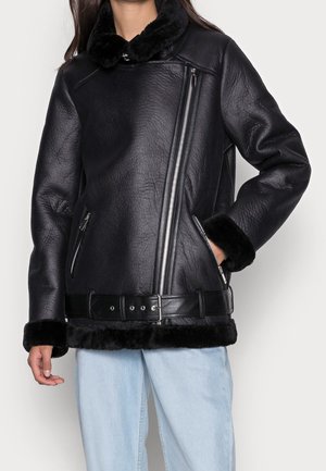 Sort læderjakke med diagonalt lynlås, pelskrave, manchetter og kant, båret sammen med lyseblå jeans. En hånd i jakkelommen.