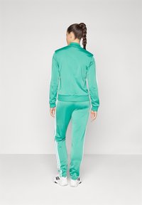Grön velourträningsdräkt med vita sidostrimlor, zip-jacka och matchande byxor; modell vänd bort, knutet hår, enfärgad design.