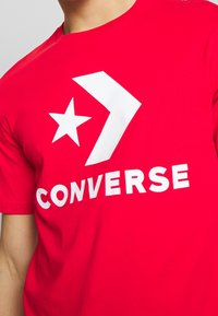 Punainen puuvillapaita, jossa on suuri valkoinen tähti ja nuoli -logo sekä sana "CONVERSE" paksuilla valkoisilla kirjaimilla.