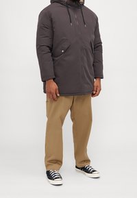 Mörkgrå vadderad jacka med huva, framdragkedja och sidofickor. Tillsammans med beige byxor och svarta sneakers med vita sulor.