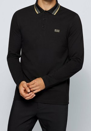 Mężczyzna noszący czarną koszulkę polo z długim rękawem, z żółto-szarym paskowanym kołnierzykiem i logo „BOSS HUGO BOSS” na lewej piersi.