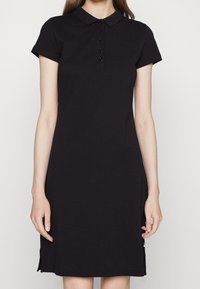 Robe polo noire à manches courtes, avec patte de boutonnage et logo brodé. Fabriquée en tissu lisse avec une coupe droite et des fentes sur les côtés.
