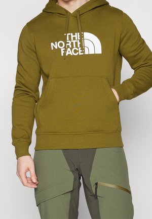 Osoba w oliwkowej bluzie z kapturem The North Face z przednią kieszenią i sznurkami, w połączeniu z dwukolorowymi zielonymi spodniami cargo z kieszeniami na zamki.