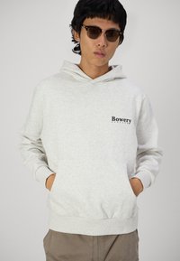 Lyst grå hættetrøje lavet af blødt stof, med en frontlomme og "Bowery" logo. Modellen bærer solbriller og har hænderne i lommerne.