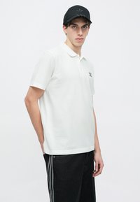 Λευκό πόλο πουκάμισο με μαύρο λογότυπο, κοντά μανίκια και κλασικό γιακά· συνδυασμένο με μαύρο παντελόνι με ρίγες στο πλάι.