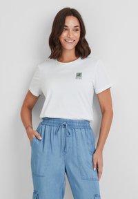 White cotton t-shirt with round neckline, featuring a green graphic print saying "RESPECT THE WILD" on the left side. Paired with light blue pants.