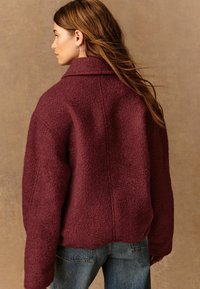 Veste oversize bordeaux en tissu texturé avec un col et des épaules tombantes, associée à un jean en denim bleu clair.