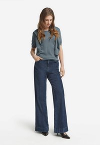 Kvinna med långt hår iförda en blå kortärmad topp, denimbyxor med vida ben, mörka spetsiga klackar och en tröja som hänger över axlarna.