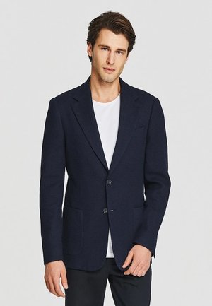 Mężczyzna ubrany w ciemnogranatową marynarkę na białej koszulce i czarnych spodniach, stoi na tle jednolitego jasnego tła.