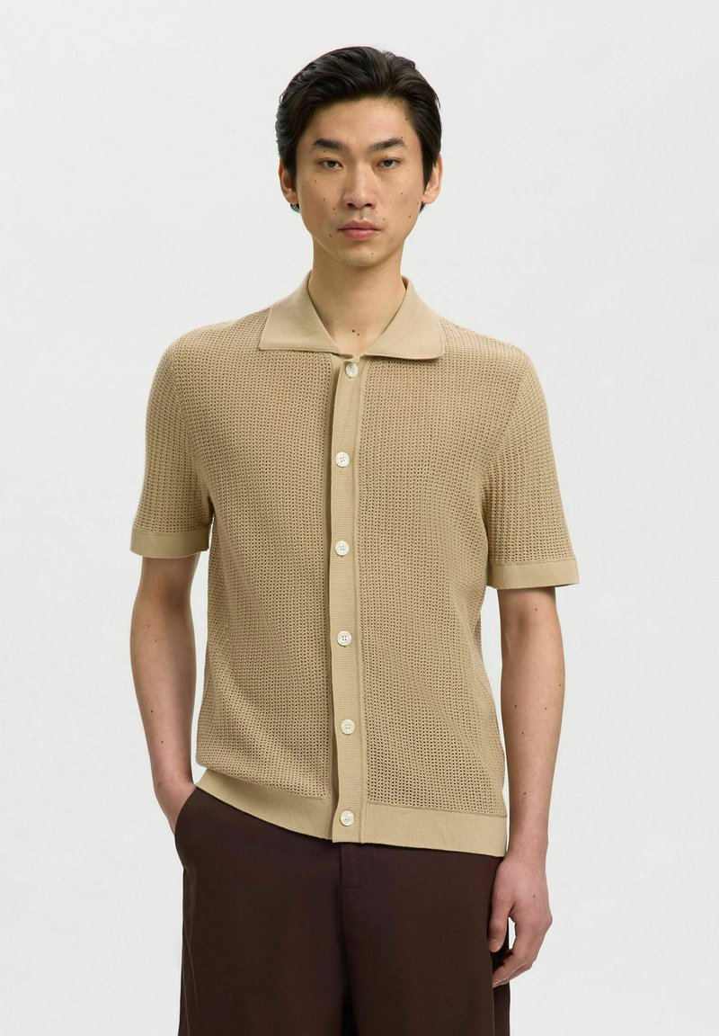 Jeune homme portant une chemise en maille beige à manches courtes avec col et un pantalon marron foncé, debout avec une main dans la poche.