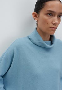 Frau trägt einen hellblauen Rollkragenpullover und silberne Creolen, sieht nach rechts mit einem neutralen Ausdruck vor einem einfarbigen Hintergrund.