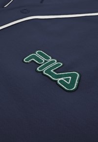 Chemise bleu marine avec bordure blanche comportant un patch logo FILA vert et blanc brodé sur le tissu.