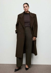 Cappotto di lana marrone doppiopetto sopra un maglione marrone e pantaloni a vita alta verde scuro, abbinati a ballerine nere a punta. Design minimalista.