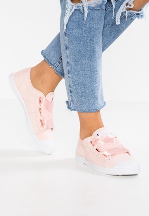 Osoba ubrana w jasnoniebieskie, podarte dżinsy oraz różowe tenisówki z szerokimi, satynowymi sznurowadłami, stojąca na białej podłodze.