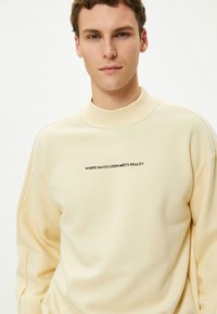 Kremowy sweatshirt z wysokim kołnierzem i luźnym krojem. Na przodzie znajduje się czarny tekst "Gdzie wyobraźnia spotyka rzeczywistość".
