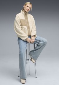 Giacca in fleece color crema con un pannello beige, colletto alto, chiusura con zip e tasca frontale. Indossata con jeans a campana azzurri chiari e scarpe beige.