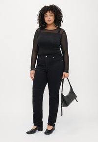 Crna majica dugih rukava s prozirnim rukavima, uparena s crnim trapericama visokog struka i crnim ravnim cipelama. Mali crni ručni rad dovršava outfit.