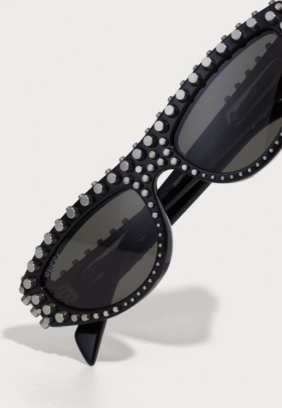 Occhiali da sole neri con montatura testurizzata, decorata con accessori in argento. Lenti ovali con tonalità scura, caratterizzate da una forma profilo laterale distintiva.