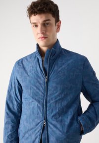 Casaco acolchoado azul com um padrão de camuflagem subtil, gola alta e fecho de correr. Apresenta bolsos laterais e um acabamento texturizado.