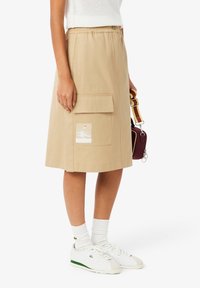 Beige knälång kjol med elastiskt midjeband, stor framficka och en glänsande lappar. Bärs med vita skor och strumpor.