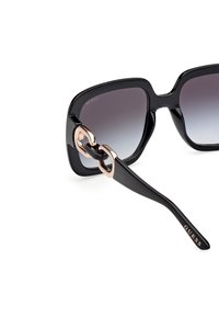 Occhiali da sole neri con montatura quadrata, lenti sfumate e un elegante accento in oro rosa lucido sulla tempia con un design intrecciato.
