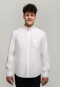 Biała koszula z długim rękawem i kołnierzykiem oraz jedną kieszonką, wykonana z gładkiego materiału. Prosty krój, noszona z ciemnymi spodniami.