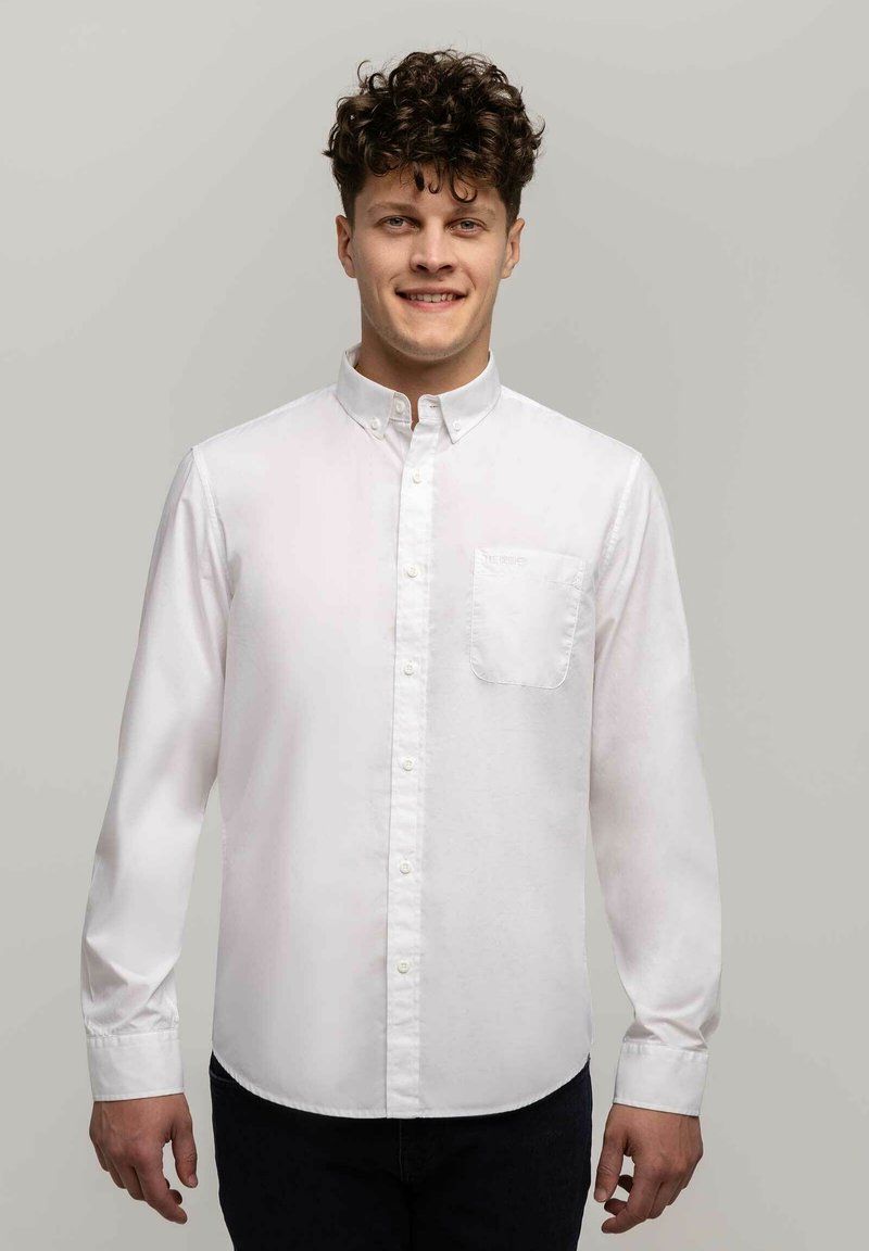 Biała koszula z długim rękawem i kołnierzykiem oraz jedną kieszonką, wykonana z gładkiego materiału. Prosty krój, noszona z ciemnymi spodniami.