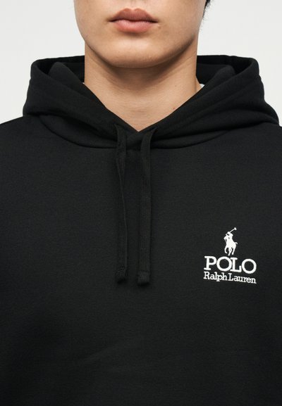 Κοντινό πλάνο ενός ατόμου που φοράει μαύρο φούτερ με κουκούλα, με το λογότυπο Polo Ralph Lauren κεντημένο σε λευκό στο στήθος.