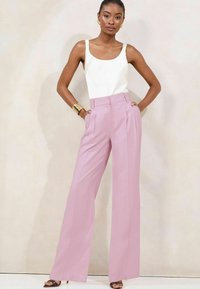 Pantaloni rosa chiaro a vita alta con pieghe e tasche laterali, abbinati a un top senza maniche bianco. Il modello indossa accessori dorati e tacchi.