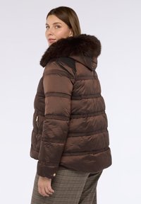 Hnědá péřová bunda s umělou kožešinou na kapuci. Má prošívaný design, zipové kapsy a žebrované detaily podél švů.