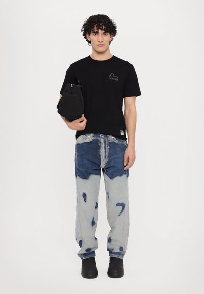 T-shirt en coton noir avec un logo, associé à un jean en denim déchiré bleu et gris, présentant des bords effilochés et des patchs. Chaussures noires.