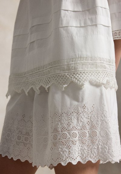 Λευκή φούστα σε στρώματα με δαντελένιο τελείωμα και περίπλοκη κέντημα με ανοιχτές τρύπες πάνω από το κυματιστό τελείωμα, εμφανίζεται σε ένα άτομο από το μέσο του μηρού έως τη μέση.