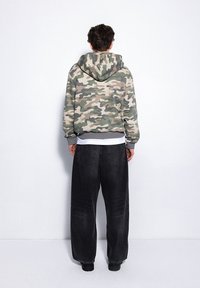 Bluza z kapturem w szaro-zielony wzór moro z sznurkiem, zestawiona z luźnymi czarnymi dżinsami i czarnymi butami.