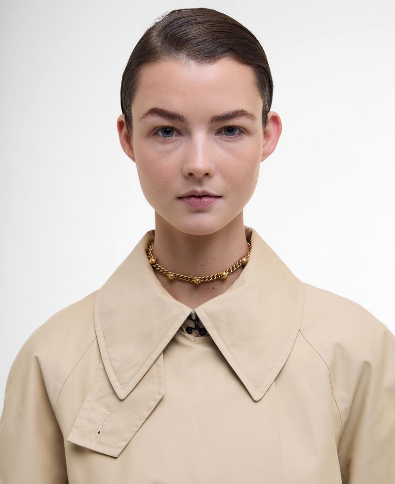 Beige Mantel mit großem Kragen, mit einer vorderen Leiste. Goldene Kettenhalskette um den Hals. Glatte Textur und minimalistisches Design.