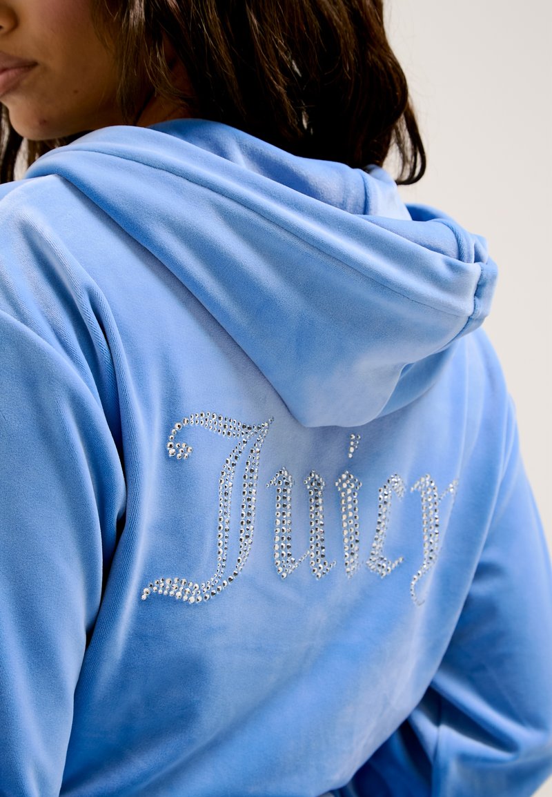 Felpa in velluto blu con vestibilità ampia, caratterizzata da testo in strass "Juicy" sul retro. Tessuto morbido e cappuccio regolabile.