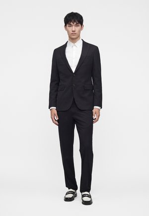 Hombre vestido con un traje negro de dos botones, camisa blanca y mocasines blancos y negros, de pie frente a un fondo blanco liso.
