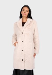 Manteau en fausse fourrure beige clair avec une texture douce, col à revers, boutons avant et poches latérales. Manches longues et design jusqu'aux genoux.