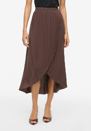 Jupe midi plissée portefeuille marron portée avec un haut noir et des talons ouverts marron, debout devant un fond clair uni.