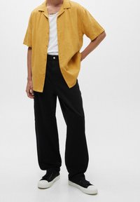 Žlutá košile s krátkými rukávy a jemným vzorem, spárovaná s černými volnými kalhotami a černými teniskami s bílými podrážkami.