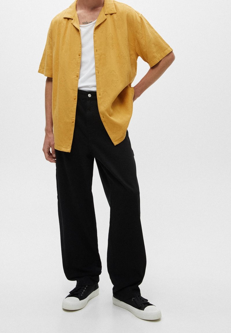 Žlutá košile s krátkými rukávy a jemným vzorem, spárovaná s černými volnými kalhotami a černými teniskami s bílými podrážkami.