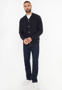 Marineblauwe cardigan met een V-hals, zes knopen, geribbelde randen en twee voorzakken, gecombineerd met donkere jeans en witte sneakers.