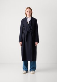 Abrigo trinchera azul marino con cintura ajustada, mangas largas y un amplio cuello, superpuesto a una camisa clara y combinado con jeans de pierna ancha.