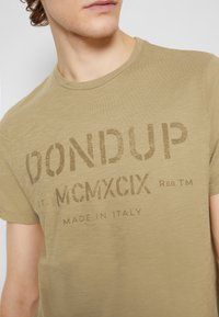 Olivgrön t-shirt i mjukt tyg, med rund hals och en stor tryckt logotyp som säger "DON DUP MCMXCIX" i mörkbrunt.