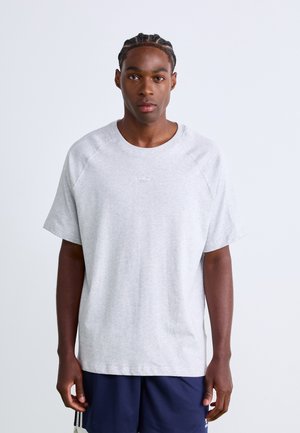 Ung man med flätat hår, klädd i en ljusgrå oversized t-shirt och marinblå sportbyxor, framför en enkel vit bakgrund.