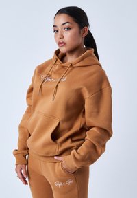 Bruine hoodie met trekkoorden, voorzak en logo. Zachte stof, relaxte pasvorm, gecombineerd met bijpassende bruine joggers met logo-detail.