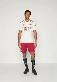 Witte voetbalshirt met rode accenten, logo "Emirates Fly Better", gecombineerd met kastanjebruine shorts, zwarte sokken met witte strepen en zwarte voetbalschoenen.