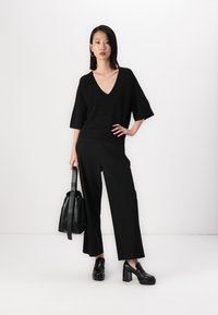 Suéter preto de malha com decote em V e mangas curtas, combinado com calças pretas de perna larga e sapatos pretos de salto grosso; a segurar uma bolsa preta.