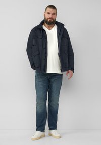 Marinejacke mit mehreren Fronttaschen, Knopfverschluss und Kapuze; kombiniert mit einem weißen V-Ausschnitt-Shirt, blauen Jeans und weißen Sneakers.
