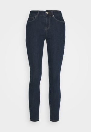 Vékony, sötétkék farmernadrág. Közepes derekú, öt zsebes kialakítással és látható varratokkal a varrás mentén.