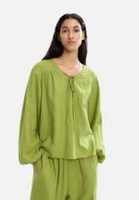 Zielona, długi rękaw bluzka z wiązaniem przy dekolcie, wykonana z miękkiej bawełny, luźny krój i marszczenia na ramionach.